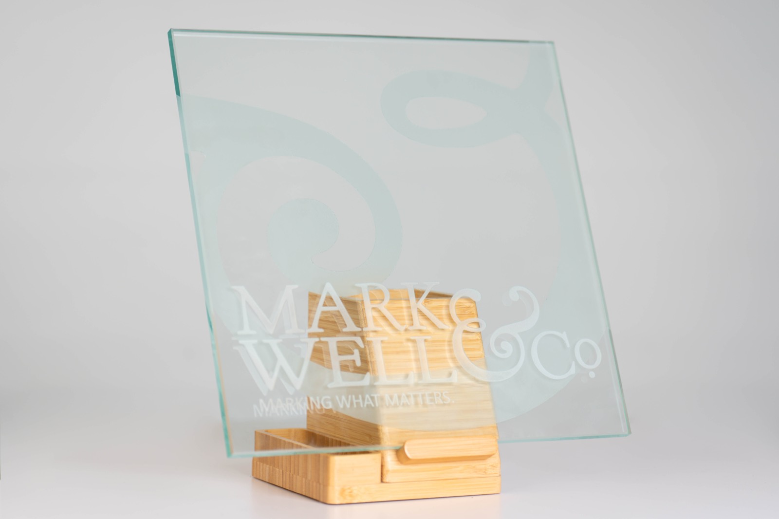 Sand-carved glass panel bearing the MarkWell & Co. mark and the line Marking What Matters, standing on a bamboo base.